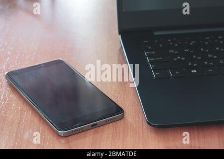 Schwarzer Laptop und Handy auf dem Tisch. Selektiver, flacher Fokus auf dem Telefon. Rücklicht. Horizontal. Stockfoto