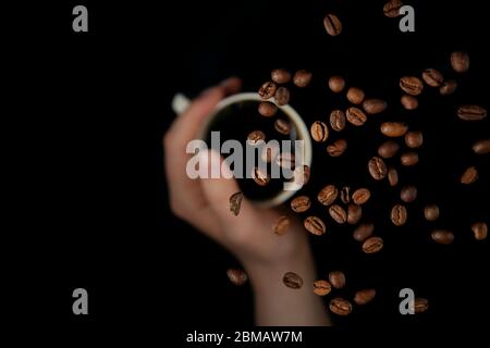 Hand halten Kaffeetasse mit schwimmenden Kaffeebohnen über, isoliert auf schwarzem Hintergrund Stockfoto