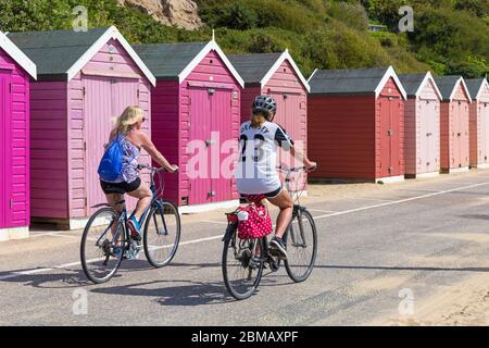 Bournemouth, Dorset, Großbritannien. Mai 2020. UK Wetter: Schöner warmer sonniger Tag mit steigenden Temperaturen am Feiertag Freitag, der VE Tag 75. Jahrestag markiert. Die Strände sind hauptsächlich verlassen, und die Leute nehmen ihre zulässige Bewegung am Meer, die meisten halten sich an die Coronavirus Richtlinien, da Besucher geraten werden, weg zu bleiben und zu Hause zu bleiben. Frauen Radfahren entlang der Promenade vorbei Strandhütten - Reiten Fahrräder Fahrrad Radfahrer Radfahrer. Quelle: Carolyn Jenkins/Alamy Live News Stockfoto