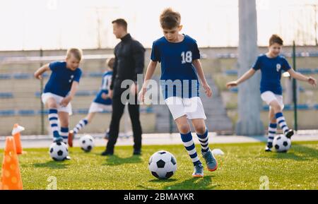 Porträt von Schuljungen Training Fußball mit jungen Trainer auf Fußballplatz. Jungen laufen mit Fußballbällen zwischen bunten Fußballmarkierungen und Kegeln Stockfoto