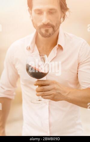 Mann mit einem Glas Rotwein. Bärtiger kaukasischer Mann in einem weißen Hemd hält ein Glas Rotwein, der darauf schaut. Weinprobe Konzept. Tonbild Stockfoto