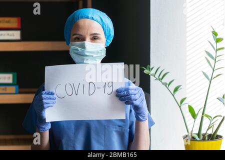 Krankenschwester oder Arzt in blauer Uniform, die ein Blatt mit der Aufschrift covid-19 hält. Junge Ärztearbeiterin steht im Büro und schaut auf die Kamera Stockfoto