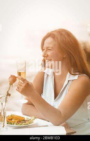 Glückliche Frau lächelt und hebt ein Glas Wein oder Champagner vor dem Hintergrund des Sandstrandes. Festlicher Toast zu Ehren eines unvergesslichen Ereignisses oder einer Hochzeit Stockfoto