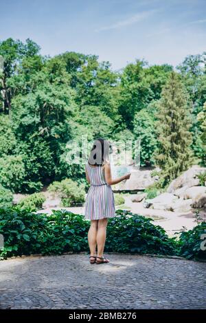 Sofia Park, Ukraine. Junges Mädchen in einem schönen Sommerpark. Mädchen in einem farbigen Sonnenkleid mit einer touristischen Karte. Frau mit einer touristischen Karte auf der Beobachtung Stockfoto