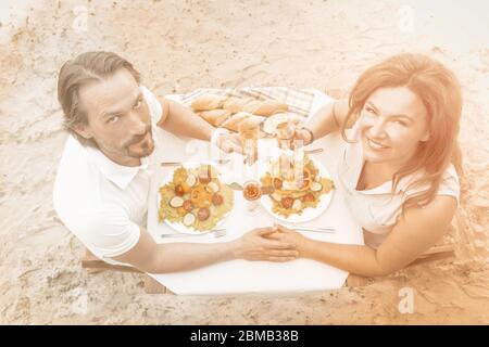 Ältere Paare haben Veranstaltung im Freien. Mann und Frau sitzen mit Nanden und heben ein Glas Wein oder Champagner vor dem Hintergrund des Sandstrandes. Hoch Stockfoto