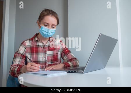 Frau in medizinischer Maske mit Laptop-Computer, Webinar ansehen, Vortrag, Notizen in grauen Raum zu Hause. Bildung, freiberuflich, Internet, Quarantäne Stockfoto