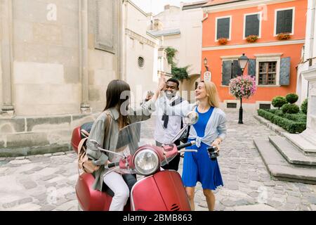 Junge drei glücklich lächelnde multirassische Menschen, dunkelhäutigen Mann und zwei kaukasischen Damen, die hohe fünf einander geben, während sie Treffen in der Altstadt Stockfoto