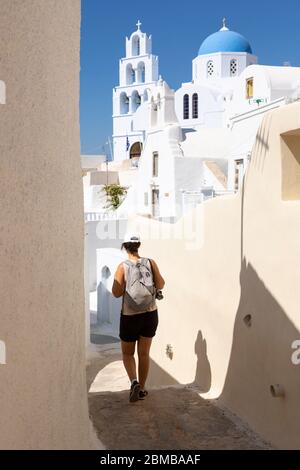 Eine Frau, die eine enge Gasse mit einer weißgetünchten griechisch-orthodoxen Kirche im Hintergrund, Pyrgos, Santorini, Griechenland, entlang geht Stockfoto