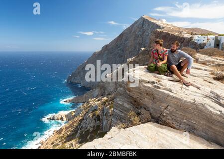 Ein junges Paar, das am Rand einer Klippe sitzt und einen Küstenblick in Folegandros, Kykladen Inseln, Griechenland genießt Stockfoto