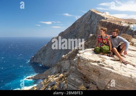 Ein junges Paar, das am Rand einer Klippe sitzt und einen Küstenblick in Folegandros, Kykladen Inseln, Griechenland genießt Stockfoto