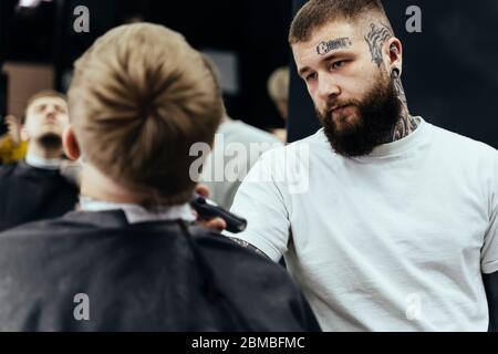 Tätowierte Barber Trimming bärtigen Mann mit Rasiermaschine im Barbershop. Frisur-Prozess. Nahaufnahme eines Frisierens, der den Bart eines Bartes schneidet Stockfoto