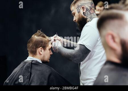 Professionelle tätowierte Friseur mit Cutthroat Rasiermesser schneiden Haar. Attraktiver Mann bekommt einen modernen Haarschnitt im Friseurladen. Nahaufnahme. Stockfoto