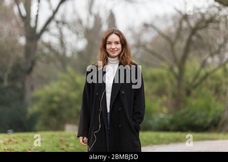 Junge Frau mit braunen Haaren und schwarzem Mantel lächelnd in Park Stockfoto