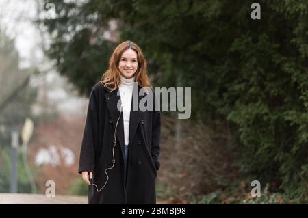 Junge Frau trägt schwarzen Mantel im Park lächelnd für Kamera Stockfoto