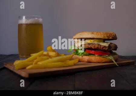 Burger und gebratene Kartoffelsorten auf einem Holzbrett und ein Bier auf seiner Seite. Alles auf einem schwarzen Holztisch und einem grauen Hintergrund. Stockfoto