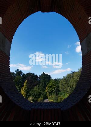 Blick auf Bäume und blauen Himmel mit Wolken durch ovale Form Steinfenster Stockfoto