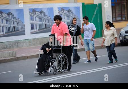 Gruppe von Asiaten, die die Straße hinunter gehen, junger Mann, der den Rollstuhl mit einer alten behinderten Frau in schwarzer Kleidung schiebt Stockfoto