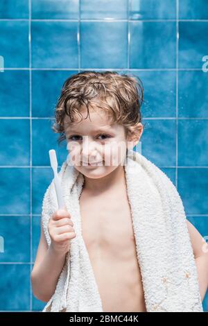 Ein Kind mit einem Handtuch auf den Schultern, das eine Zahnbürste zeigt, mit blauen Fliesen im Hintergrund. Stockfoto