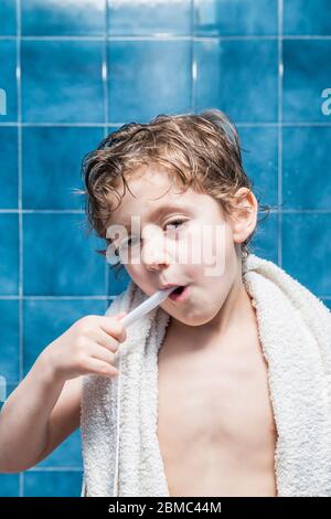 Ein Kind mit einem Handtuch auf den Schultern, das seine Zähne putzt, mit blauen Fliesen im Hintergrund. Stockfoto