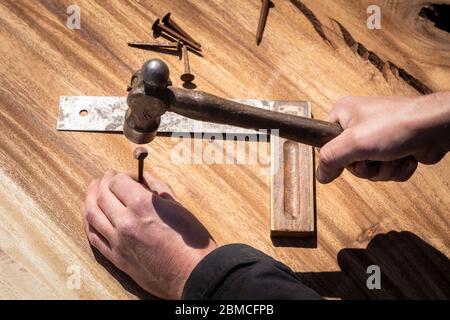 Hand eines Zimmermanns, der einen Kugelhammerschlag hält, um einen rostigen alten Nagel in ein Stück Holz zu treiben. Messwerkzeug auf dem Hintergrund. DIY, authentische Maki Stockfoto