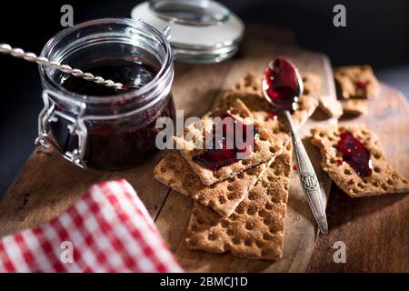 Offenes Glas mit Johannisbeer-Marmelade auf einem Holzbrett. Trockene Kekse mit Marmelade, Krümel und einem Löffel in Marmelade bestäuben. Platz für Text. Stockfoto