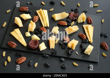 Stücke von Parmesan und gouda mit Bockshornklee, blauen und goldenen Rosinen und getrockneten Aprikosen auf schwarzem Steinteller auf schwarzem Tisch. Nahaufnahme Stockfoto