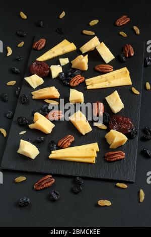 Stücke von Parmesan und gouda mit Bockshornklee, blauen und goldenen Rosinen und getrockneten Aprikosen auf schwarzem Steinteller auf schwarzem Tisch. Nahaufnahme Stockfoto