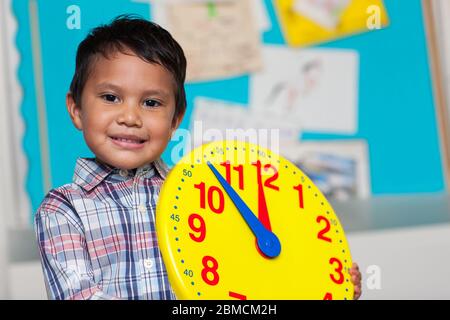 Kleiner Junge mit einem preppy Hemd, hält eine große analoge Uhr mit einem bunten Stierkampfbrett im Hintergrund. Stockfoto