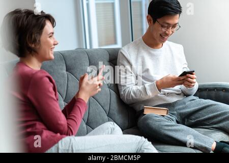 Foto von fröhlichen multinationalen Paar lachen und mit Smartphones sitzen auf dem Sofa zu Hause Stockfoto