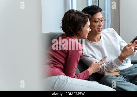 Foto von fröhlichen multinationalen Paar lachen und mit Smartphones sitzen auf dem Sofa zu Hause Stockfoto