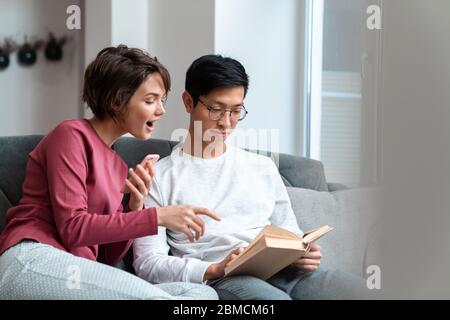Foto von begeisterten multinationalen Paar Buch lesen und mit Smartphone während auf dem Sofa zu Hause sitzen Stockfoto