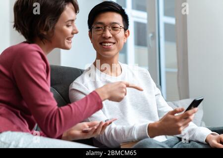 Foto von fröhlichen multinationalen Paar mit dem Finger auf das Smartphone während des Sitzens auf dem Sofa Stockfoto
