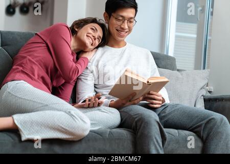 Foto von lächelnden multinationalen Paar Buch lesen und mit dem Smartphone sitzen auf dem Sofa zu Hause Stockfoto