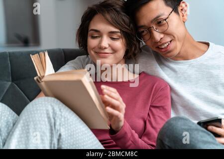 Foto eines lächelnden multinationalen Paares, das Buch liest und das Handy benutzt, während es zu Hause auf dem Sofa sitzt Stockfoto