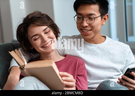 Foto eines lächelnden multinationalen Paares, das Buch liest und das Handy benutzt, während es zu Hause auf dem Sofa sitzt Stockfoto