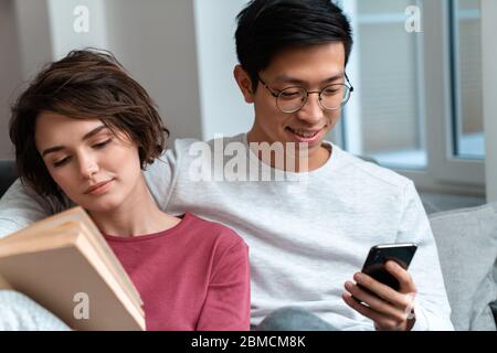 Foto von einem multinationalen Paar, das das Buch liest und Smartphone benutzt, während es zu Hause auf dem Sofa sitzt Stockfoto