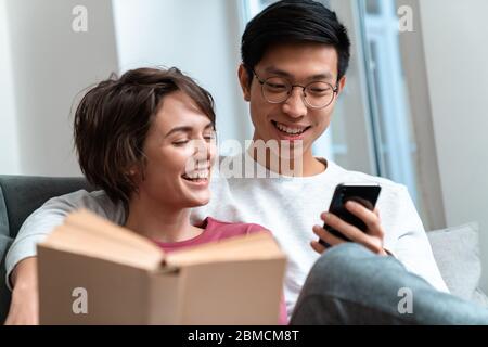 Foto von lachenden multinationalen Paar Buch lesen und mit Handy während sitzen auf dem Sofa zu Hause Stockfoto