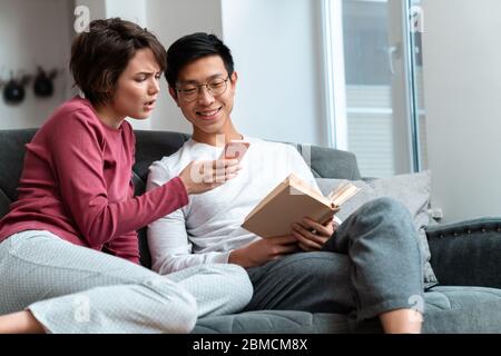 Foto von schockierten multinationalen Paar Buch lesen und mit Smartphone während auf dem Sofa zu Hause sitzen Stockfoto