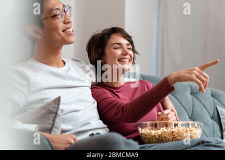 Foto von fröhlichen multinationalen Paar Popcorn essen und Filme sehen, während auf dem Sofa zu Hause sitzen Stockfoto