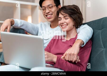 Foto von fröhlichen multinationalen Paar lachen und Laptop während sitzen auf dem Sofa zu Hause Stockfoto
