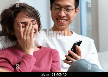 Foto von fröhlichen multinationalen Paar lachen und Handy während sitzen auf dem Sofa zu Hause Stockfoto