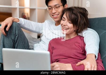 Foto von fröhlichen multinationalen Paar lachen und Laptop während sitzen auf dem Sofa zu Hause Stockfoto