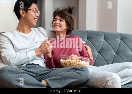 Foto von fröhlichen multinationalen Paar Popcorn essen und einander anschauen, während sie auf dem Sofa zu Hause sitzen Stockfoto
