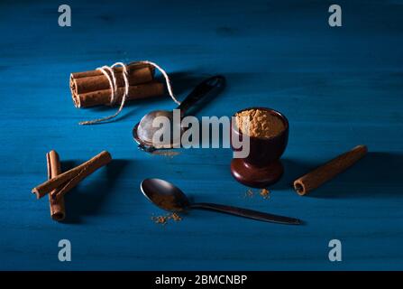 Gemahlenes Zimtpulver in Holzbecher, eine Packung Zimtstangen, ein Löffel und Metallsieb auf blauem Holztisch mit dunklem Hintergrund. Ceylon cin Stockfoto