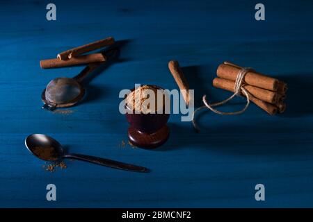 Gemahlenes Zimtpulver in Holzbecher, eine Packung Zimtstangen, ein Löffel und Metallsieb auf blauem Holztisch mit dunklem Hintergrund. Ceylon cin Stockfoto