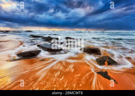 Sandstrand mit dunklen Sandsteinfelsen bei Sonnenaufgang an den nördlichen Stränden des Pazifischen Ozeans von Sydney. Stockfoto