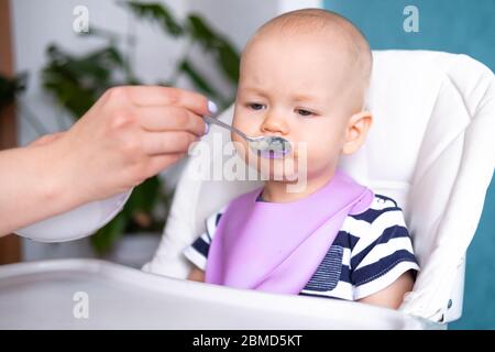 Trauriges Baby, das geschmackloses Essen isst. Mama füttert kleines kaukasisches Kind auf Hochstuhl. Gesunder Brei, Mutter Hände mit Löffel. Neugeborene Kinder Stockfoto