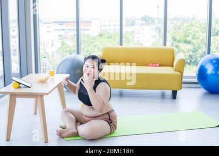 Asiatische übergewichtige Frau mit Tablette und. Tragen Kopfhörer für Musik hören und sie ist die Ausübung zu Hause, asiatische Mädchen genießen Essen und Popcorn Stockfoto