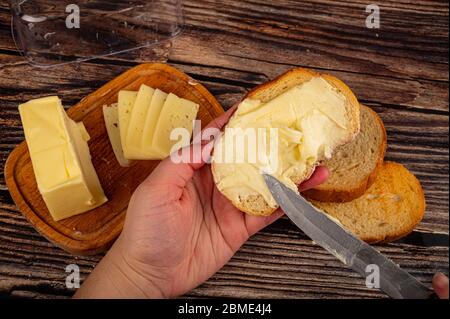 Jemand breitet ein wenig Butter mit einem Messer auf frischen Weizenstoast, ein Stück Butter in einer hölzernen Butterdose und frischen Weizen Toast auf einem hölzernen Backgr aus Stockfoto