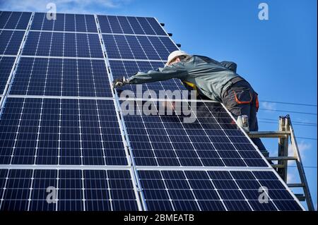 Professioneller Elektriker auf Leiter stehen und Installation von Photovoltaik-Solarpanel-System. Mann Techniker im Schutzhelm unter blauem Himmel. Konzept der alternativen Energie und Energie Stockfoto